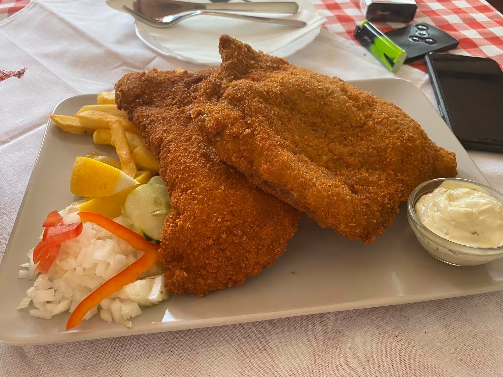 Breaded fried fish fillets served with French fries, lemon wedge, chopped onions, cucumber slices, red bell pepper, and a side of tartar sauce on a white plate.