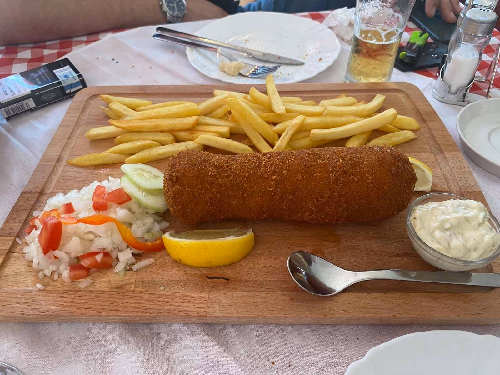 Wooden board with breaded fried croquette, French fries, sliced cucumber, diced onions, red and orange bell pepper, lemon wedge, and a small bowl of creamy sauce with a spoon.