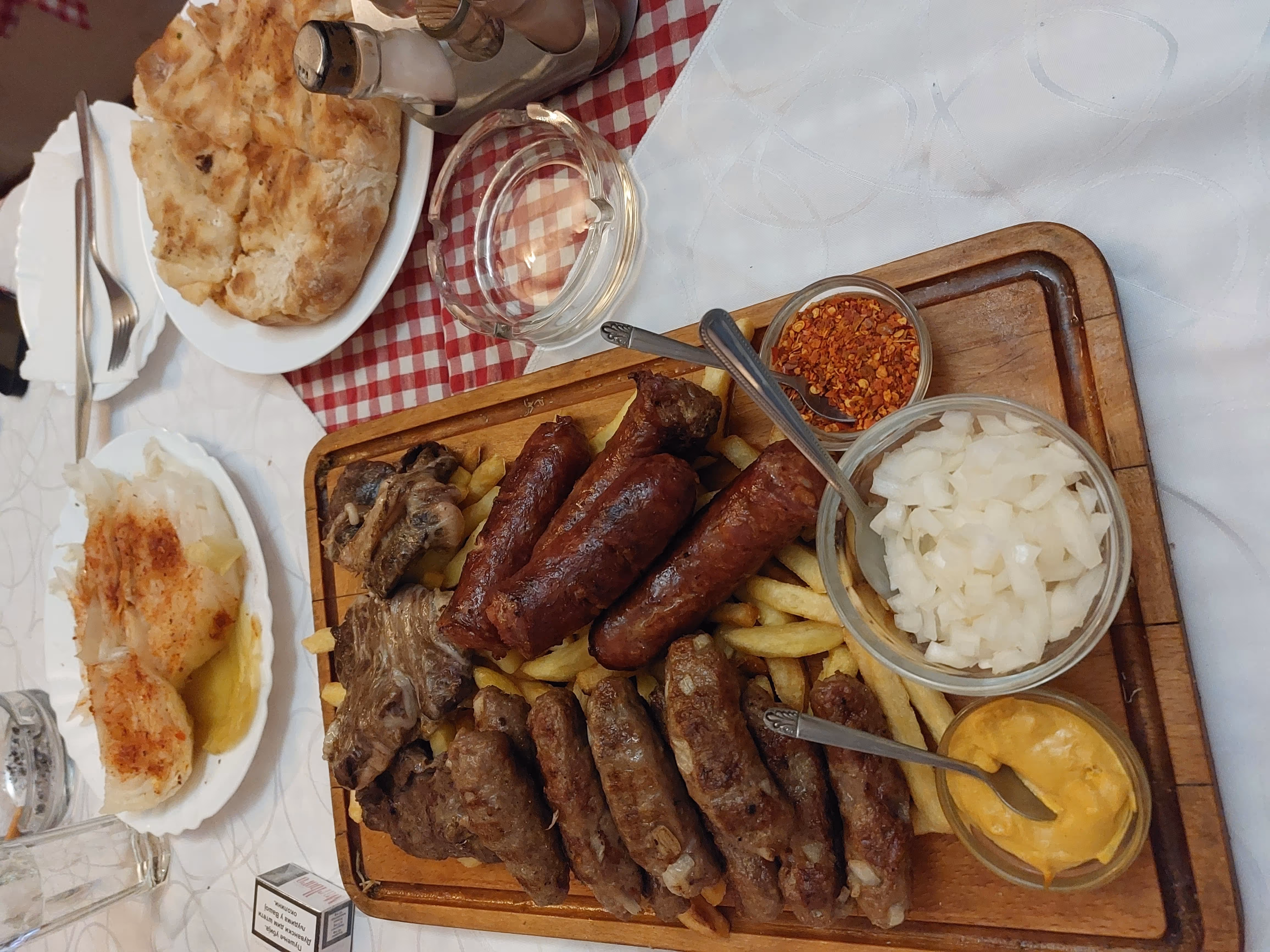 Wooden board with grilled sausages, meat, fries, bowls of chopped onions, mustard sauce, and chili flakes, accompanied by flatbread and a plate with grilled fish on a table.