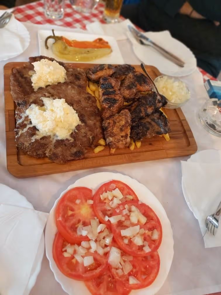 Wooden board with grilled meat patties topped with white cheese and grilled chicken pieces on fries, served with a plate of sliced tomatoes and chopped onions.