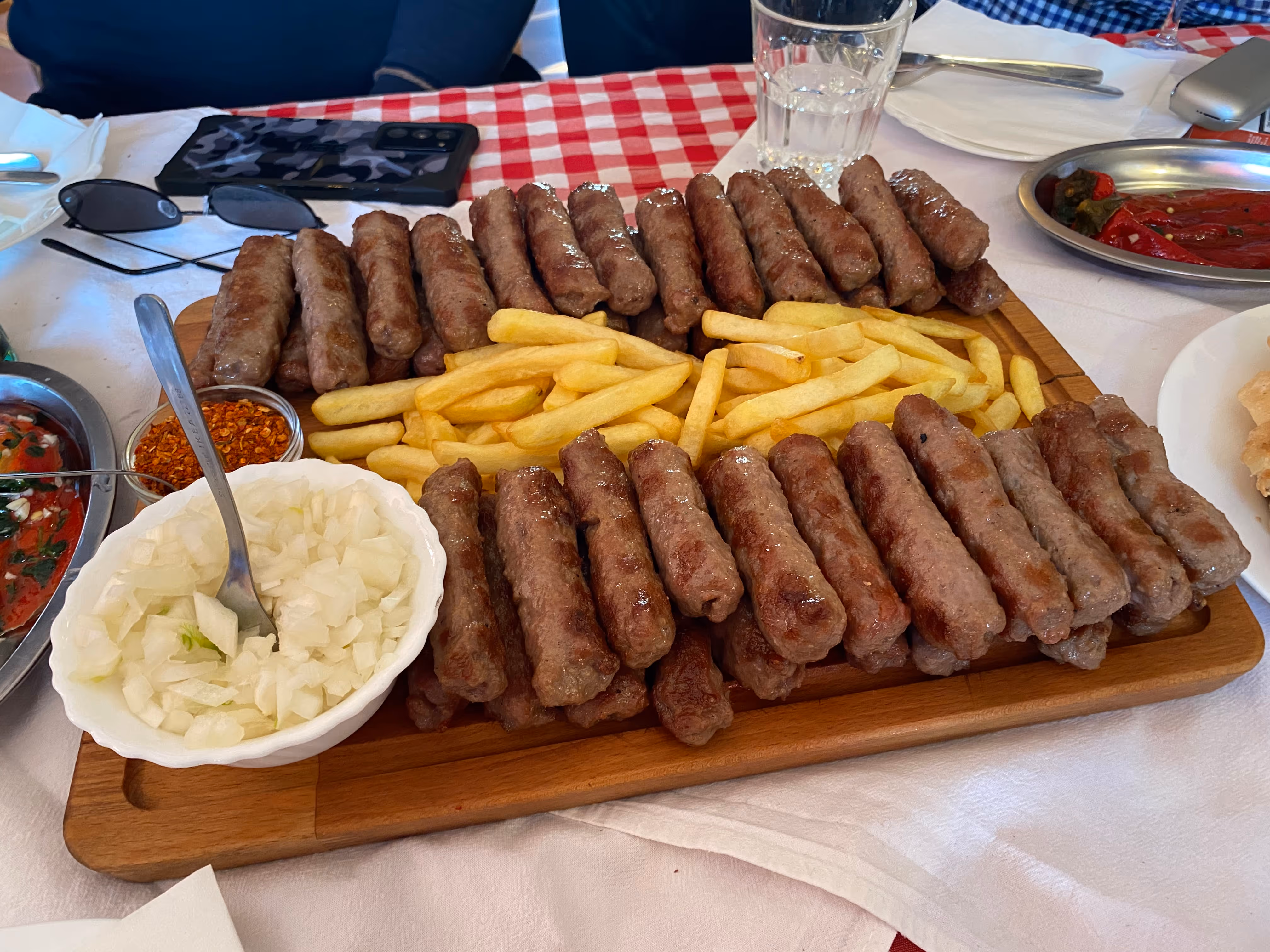 Wooden board with grilled sausages and French fries, accompanied by a bowl of chopped onions and a small container of red chili flakes on a table with a red and white checkered tablecloth.