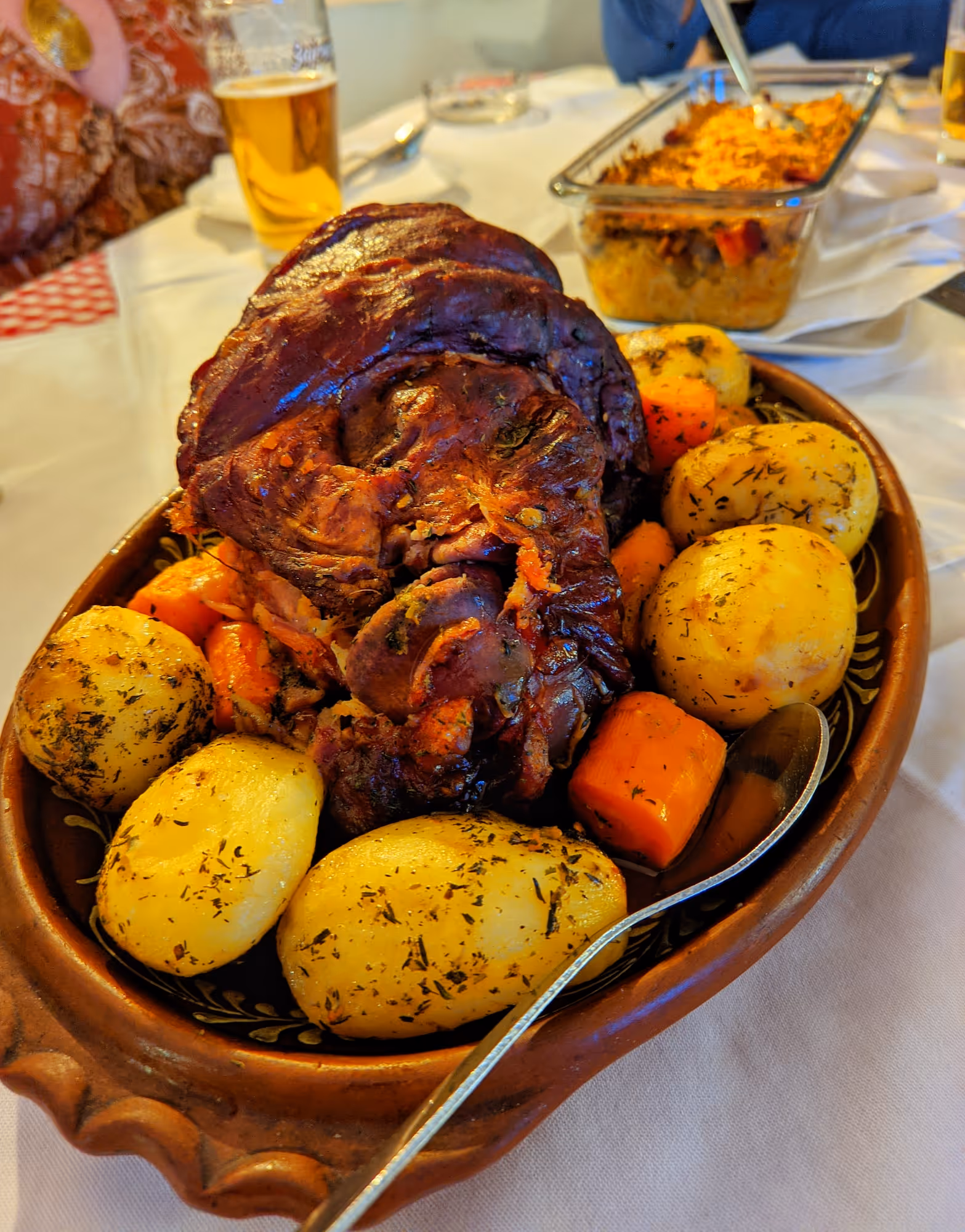 Roasted meat served with herb-seasoned potatoes and carrots in a brown ceramic dish on a white tablecloth.