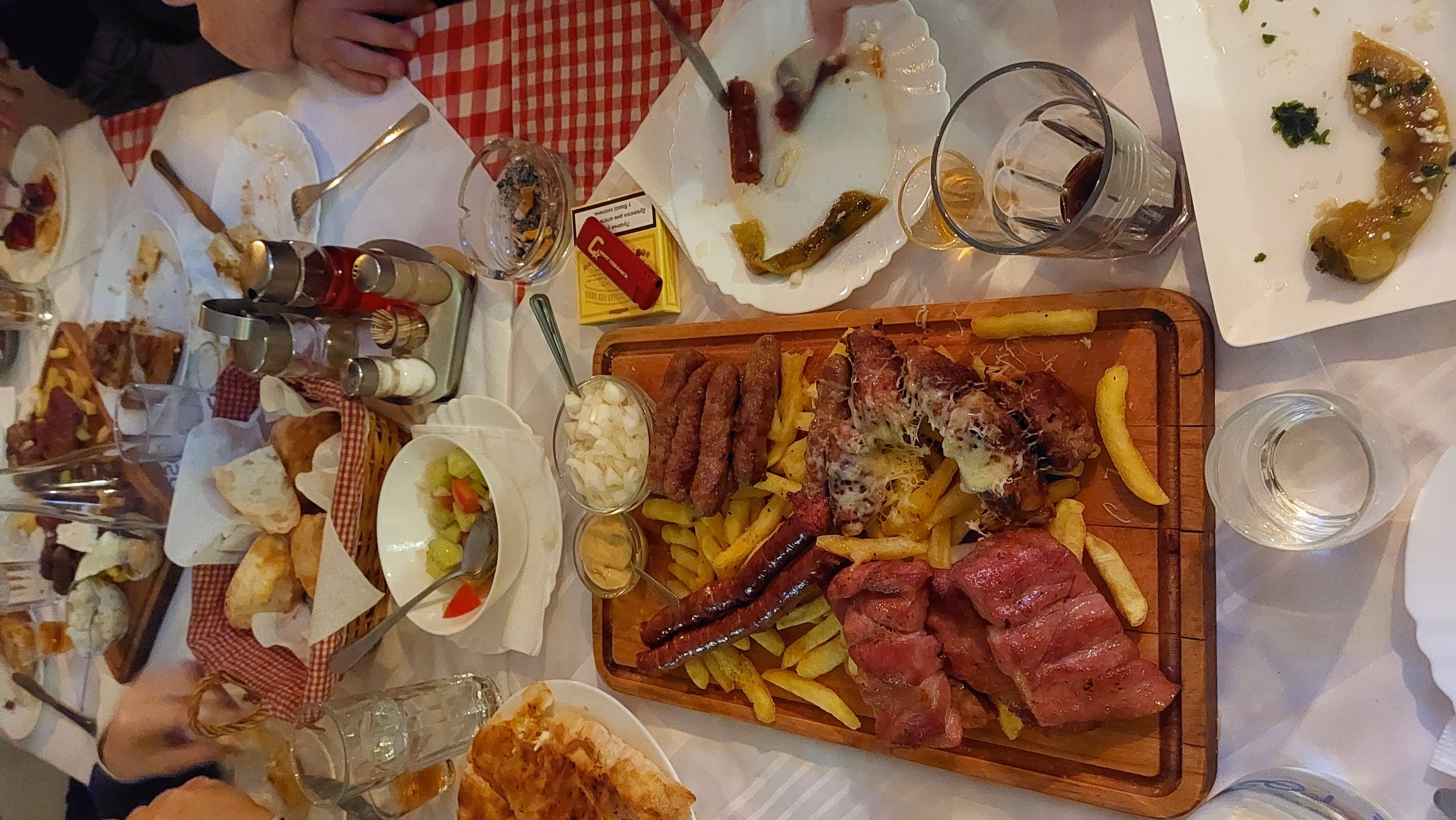 Table setting with a wooden board of grilled meats and fries, a basket of bread, various condiments, drinks, and partially eaten plates.