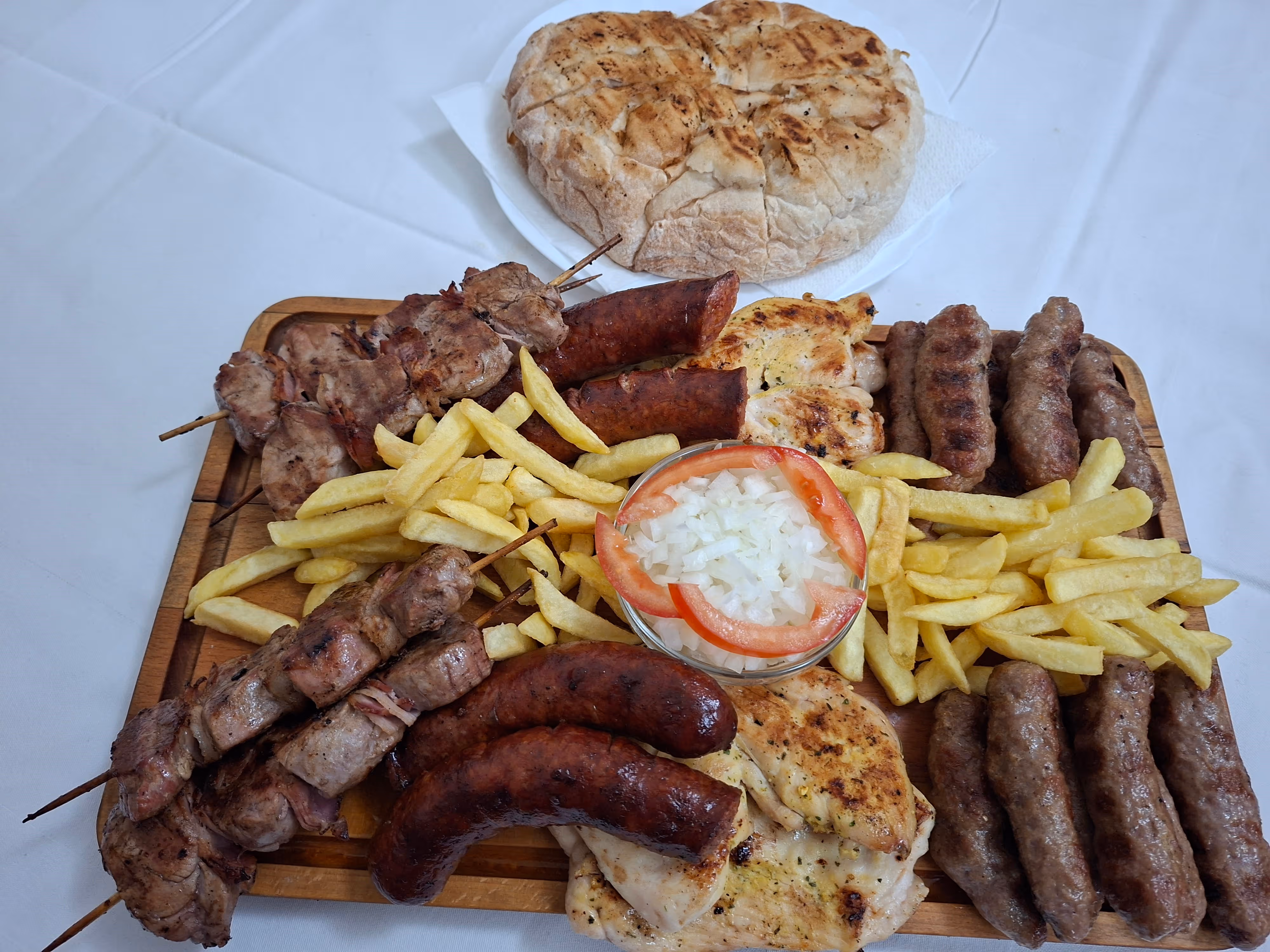 Plate with two grilled sausages, French fries, sliced onions with herbs, and a small bowl of mustard, with a bread roll on a separate plate in the background.